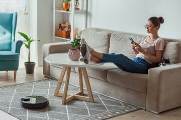 Robotic vacuum cleaner cleaning carpet, woman remote control mobile phone and enjoy rest, sitting...