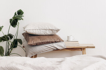 Fototapeta na wymiar Trendy Scandinavian interior still life. Velvet and linen cushions on vintage wooden bench, table. Cup of coffee on pile of books and monstera potted plant. White wall background. Blurred bed