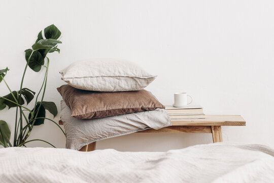 Trendy Scandinavian interior still life. Velvet and linen cushions on vintage wooden bench, table. Cup of coffee on pile of books and monstera potted plant. White wall background. Blurred bed