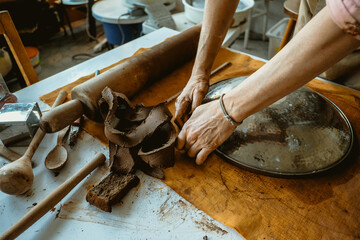A woman in an apron works with her hands with clay. On the table clay, rolling pin, piece of cloth and metal bowl	