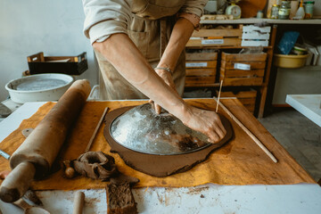 A woman in an apron works with her hands with clay. On the table clay, rolling pin, piece of cloth and metal bowl	