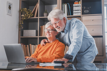 Happy elderly 60s couple sit rest on couch at home pay household expenses online on computer,...