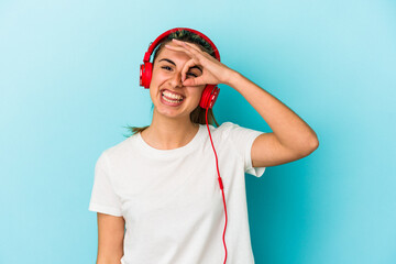 Young blonde woman listening to music on headphones isolated on blue background excited keeping ok gesture on eye.