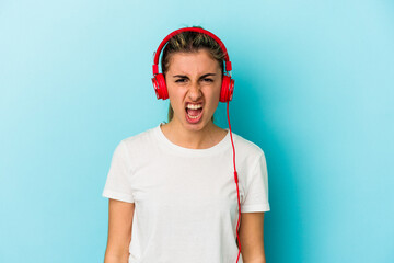 Young blonde woman listening to music on headphones isolated on blue background screaming very angry and aggressive.