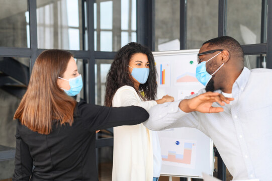 Diverse two women and man, colleagues in protective face masks greeting bumping elbows at workplace, woman coworkers in facial covers protect from COVID-19 coronavirus in office, healthcare concept