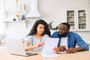 Puzzled thoughtful multiracial man and woman sitting together on table, studying documents of their...