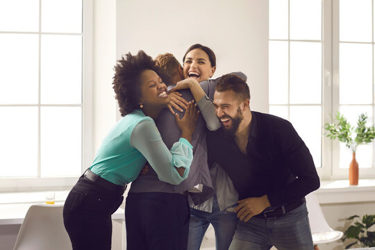 Group Of Overjoyed Young Diverse People Hugging Each Other. Team Of Excited Office Workers And Friends Embracing Their Colleague Congratulating Him On Promotion Or Happy Life Event