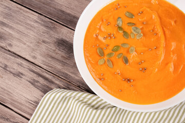Top view on pumpkin cream soup in white bowl toped with pumpkin seeds on wooden table background with copy space for text. Vegan food concept, homemade soup recipe. Selective focus