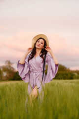 Stylish girl in violet dress and hat in field. Happy young woman on the sunset or sunrise in summer nature. Fashion and lifestyle concept.