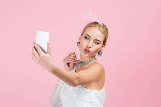 blonde woman holding paper crown on stick and taking selfie while sending air kiss isolated on pink