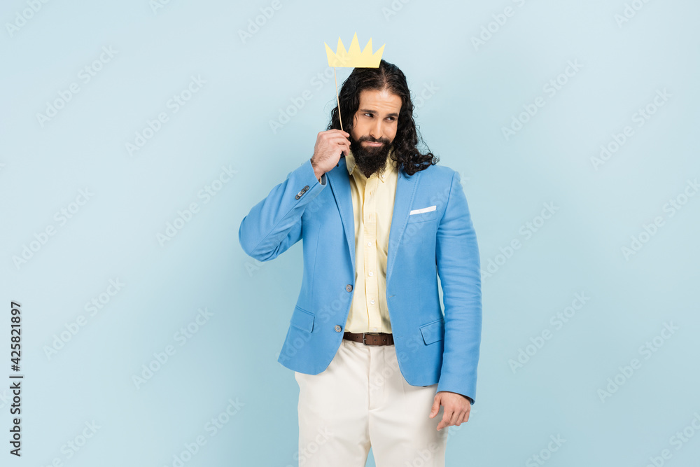 Wall mural sad hispanic man in jacket holding paper crown on stick isolated on blue