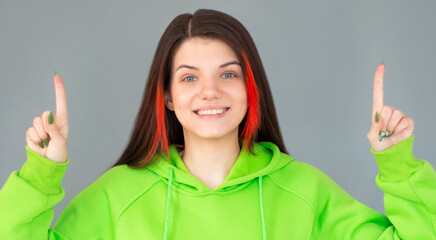 Photo of cheerful happy woman dressed in green hoodie pointing up over grey background. Look at camera.