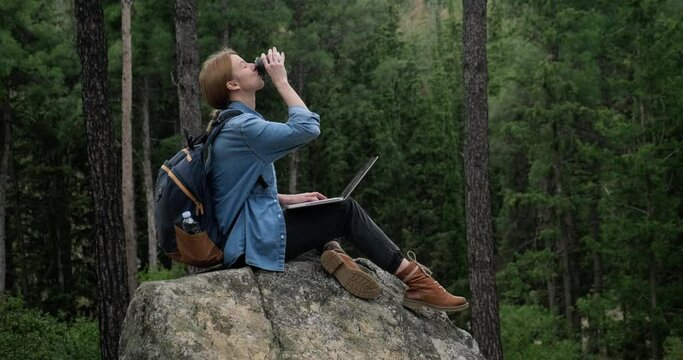 Young freelancer woman with backpack working on the laptop in the forest sitting on huge rock.