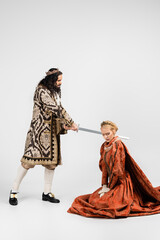 full length of cruel hispanic king in medieval clothing holding sword near scared blonde queen in crown sitting on white