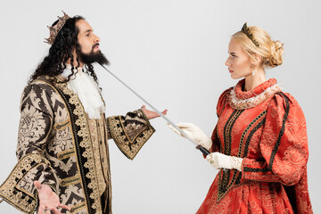 blonde queen holding sword near neck of hispanic king in medieval clothing and crown isolated on...