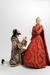 hispanic king in crown and medieval clothing standing on knee while blonde queen in dress showing no gesture on white