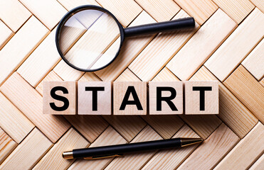 Wooden cubes with the word START stand on a wooden background between a magnifying glass and a pen