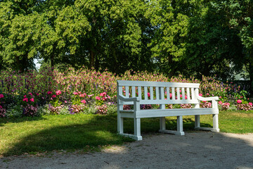 A white park bench in front of large colorful flowerbed in sunlight