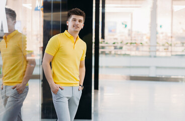 Portrait of a handsome smiling young man in a yellow polo and light trousers near a shop window