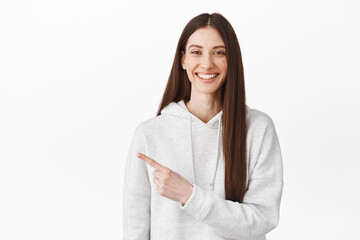 Follow this way, visit link. Smiling female student pointing finger aside. Girl showing advertisement, indicating at logo on copy space, standing over white background