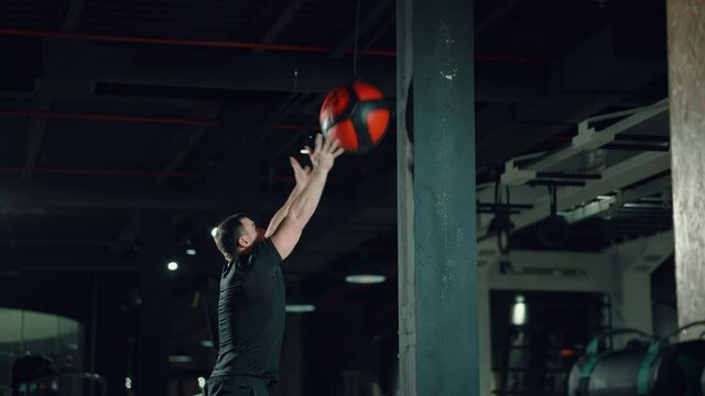Man in black sportswear exercising with medicine ball in dark gym, throwing it up and squatting. Muscular athlete doing high intensity interval training. Concept of sport