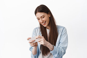 Happy young woman playing video game on smartphone, laughing and looking at phone screen, enjoying gaming, standing against white background