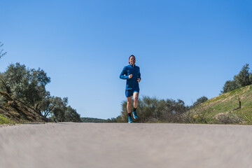 A young white european and athletic man is running in the countryside on a sunny day. The guy is wearing blue clothes and a cap.