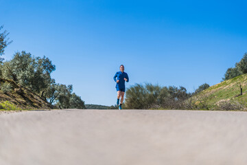 A young white european and athletic man is running in the countryside on a sunny day. The guy is wearing blue clothes and a cap.