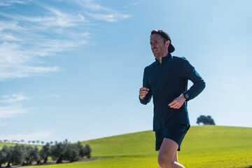 A young white european and athletic man is running in the countryside on a sunny day. The guy is wearing blue clothes and a cap.