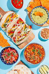 Mexican food table with traditional dishes. Chili con carne, tacos, tomato salsa, corn chips with guacamole. Mexican feast in hard light on blue color background, top view