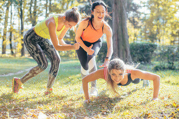 Fototapeta premium Fitness group working out at park. Team of three slim women outdoor doing fitness exercise. Laughing girls counting for their friend during plank exercise in park.