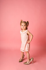 little girl in a pink lace dress and her mother's shoes stands on a pink background with space for text