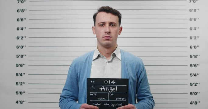 Portrait of man in shirt and cardigan holding sign for photo in police department. Crop view of male person in 30s posing, raising head and looking to camera. Concept of mugshot and crime.