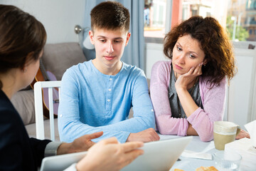 Focused teenage boy and his mother sitting at home table, attentively listening college tutor