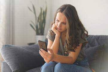 Bored young woman spends leisure time in social networks, holds smartphone. Depressed melancholy female with a mobile phone sitting on the couch at home