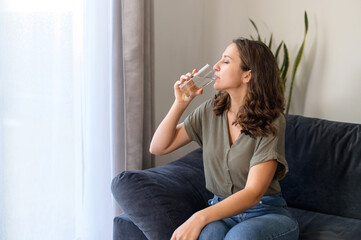 Young curly woman feels thirst at home, drinking pure fresh water from transparent glass, restores...