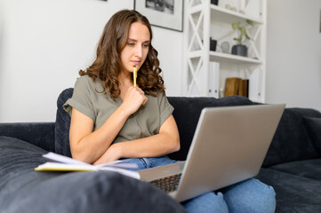 Thoughtful young woman with a laptop, looking on the screen concentrated, takes a notes. Female...
