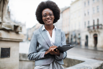 Front view of smiling african business woman in stylish formal clothes holding clipboard on city...