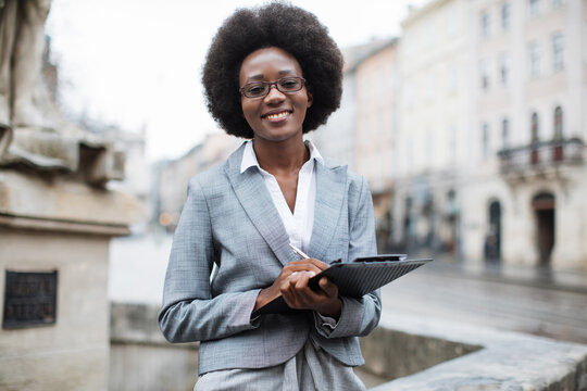 Front view of smiling african business woman in stylish formal clothes holding clipboard on city street. Concept of successful career.
