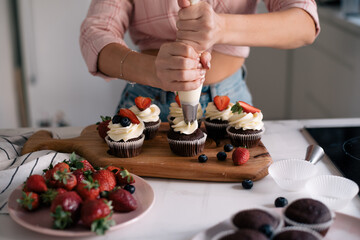 Process of making cupcake by hand.  Baking cake in the kitchen chocolate muffin on wooden cut board with strawberry, cream and blueberry. Woman’s hands push cream from pastry silicone bag.