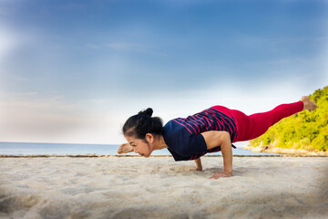Strong asian senior woman do one legged arm balance Hurdler yoga pose at the sandy beach with blue sky and beautiful sea in the summer of Phuket, Thailand. Sport active lifestyle and healthy concept.