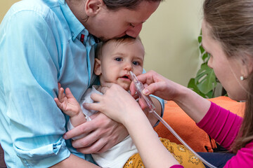 Father holding and kissing his baby boy while the mother is using the nasal aspirator to clear the mucus from the nose of her infant. Baby healthcare concept.