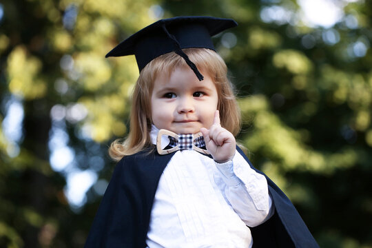 Baby in mantle outdoor. Little boy child in black academic gown and squared school hat and bow tie standing outdoor on green natural background. - Powered by Adobe