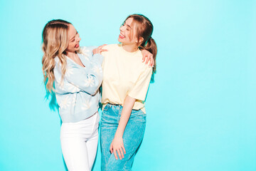 Two young beautiful blond smiling hipster female in trendy summer clothes. Sexy carefree women posing near blue wall in studio. Trendy and positive models having fun