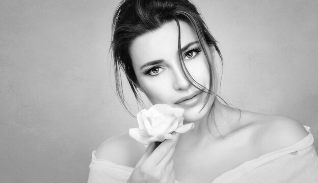 Monochrome beauty portrait of a beautiful woman with a flawless healthy skin holding a fresh white rose to face. Youth and skin care concept