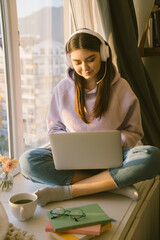 Fototapeta premium Smiling woman with headphones working online with laptop. Girl freelancer sitting near window on the sunset at home. Distance learning and remote job concept.