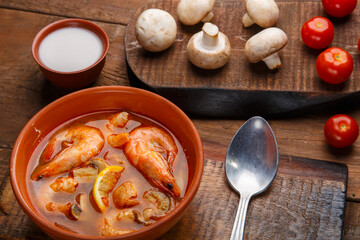 Tom yam soup with shrimps in a plate on a wooden table with a spoon next to a bowl with coconut milk mushrooms and tomatoes.
