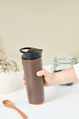 Woman brews tea in a brown thermo mug to take with her. She is holding a cute wooden spoon in the is background of a glass jar with loose tea. Healthy food concept. Reusable eco-friendly kitchen