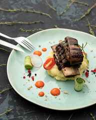 Fried rib meat on pillow of mashed potatoes decorated with fresh cucumber rolls and tomato. Delicious restaurant serving dishes on black table with cutlery, fork and knife on it. Close up shot.