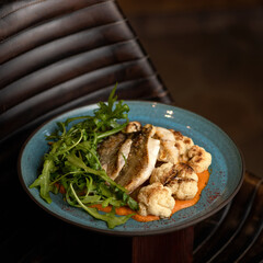 Hearty omega-3 diet lunch. Fried river fish with vegetables. Food plate on black background. Cooked Fish fillet with cauliflower and arugula salad. Close up shot. 